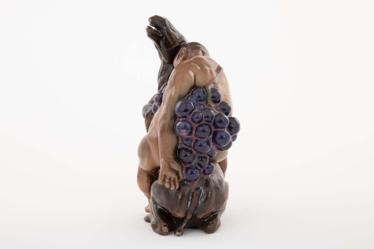 Figurgruppe i stentøy med en stor, sovende mannsfigur, sittende på et esel, drueklaser, og en liten figur som henger i eselets ører. Den lille fiiguren er en faun eller satyr, den store er antagelig en fremstilling av Bacchus. Figurgruppen er brunglasert i lyse og mørke nyanser, samt blått og fiolett.