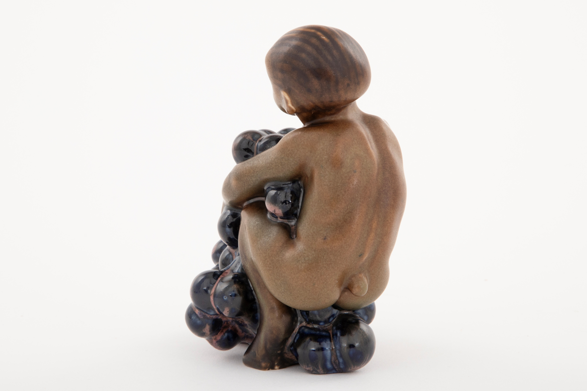 Figur i stentøy; en ung faun med drueklaser. Figuren er brunglasert i lyse og mørke varianter, samt blått og fiolett.