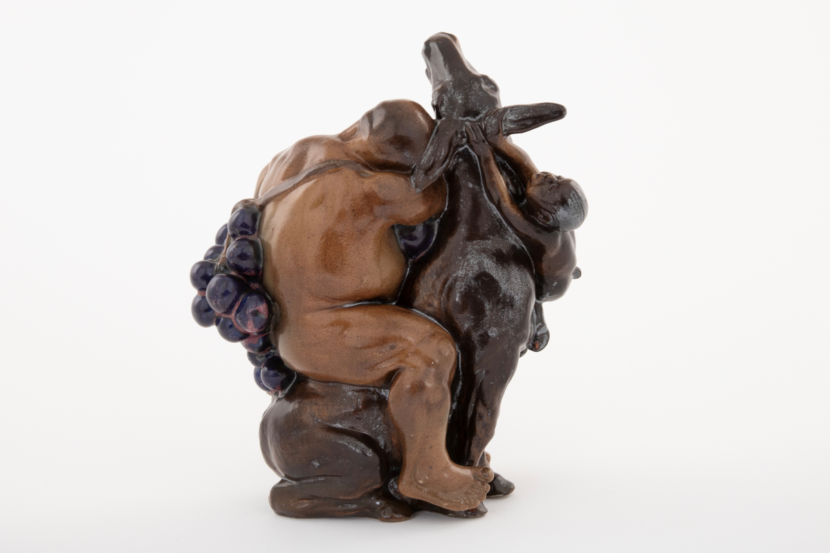 Figurgruppe i stentøy med en stor, sovende mannsfigur, sittende på et esel, drueklaser, og en liten figur som henger i eselets ører. Den lille fiiguren er en faun eller satyr, den store er antagelig en fremstilling av Bacchus. Figurgruppen er brunglasert i lyse og mørke nyanser, samt blått og fiolett.