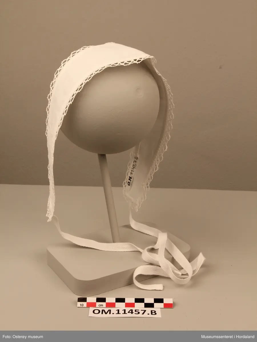 Langt og smalt hårbånd av hvit bomull med spiss på den ene langsiden. Den har maskinbrodert kant og knytebånd.