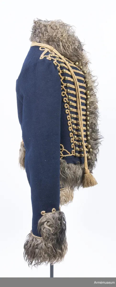 Blå päls med krage och ärmuppslag av gråbrunt fårskinn, med snören av gult ullgarn.

Pälsen är sydd av mörkblått kläde och är helfodrad med vitt, numera gulnat, korthårigt fårskinn. Ärmar, krage, framkanter och nederkant har bräm av gråbrunt fårskinn med lång lugg.
Plagget är konstruerat med fram- och sidstycken i ett samt helt bakstycke med litet skört i sidsömmarna. Ärmarna är ensömsärmar eller S-ärmar.
Tvärs över bröstet löper 18 rader snörmakerier gjorda av gul löst spunnen tvåtrådig ullsnodd. 5 knapprader, varav de två yttersta på varje sida har kullriga knappar medan knappraden i mitten som är avsedd att knäppas har klotknappar. Samtliga knappar är gjorda i en mässingslegering.
Ärmarna är kantade med päls. Pälsbrämet går upp i en "ruta" på framärmen, kantad med snodd. På baksidan av plagget nertill under ärmarna sitter två separata "rutor" av päls kantad med snodd. Sidsömmarna bak är markerade med snoddar som också pryder skörtets veck.
På baksidan av plagget nedanför kragen är det fäst ett flätat hängsle vars ändar hänger ner från varje axel och fram till plaggets främre nederkant. Varje del av hängslet är avslutat i en tofs. Vänster hängsle har ett spolformat knäppe klädd med snörmakeriarbete, medan det högra har en knut. Dessa är avsedda att hålla ihop plagget när det hänger öppet över vänster axel.
På vänster axel sitter en axelträns av dubbel snodd som sätts fast med en mässingsknapp.
