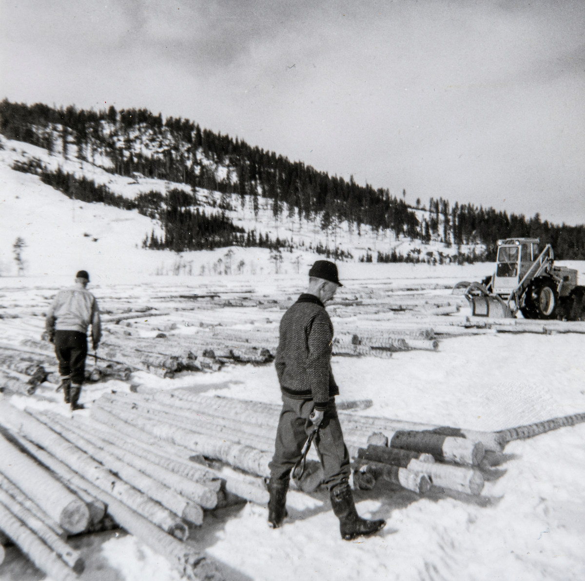 Åstadalen
To tømmermålere i arbeid med tømmeret. Lastemaskinen er i ferd med å skyve sammen tømmeret for å slippe det i elva for fløting. Tømmeret er blitt maskinbarket.