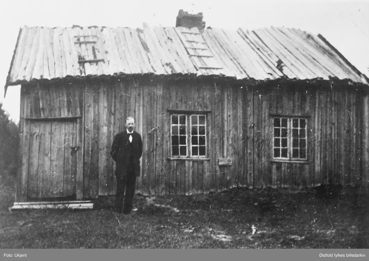 Haugen, husmannsplass under Trandem søndre i Trøgstad ca. 1912-15.
Plassen lå mellom Stikla og Lundhagen. Fraflyttet i 1912, revet ca. 1920. Mannen som står utenfor er sønn av siste bruker, Bernhard Haugen. På et annet fotokort med samme motiv er det opplyst at personen på bildet er Bernhard Haugen.