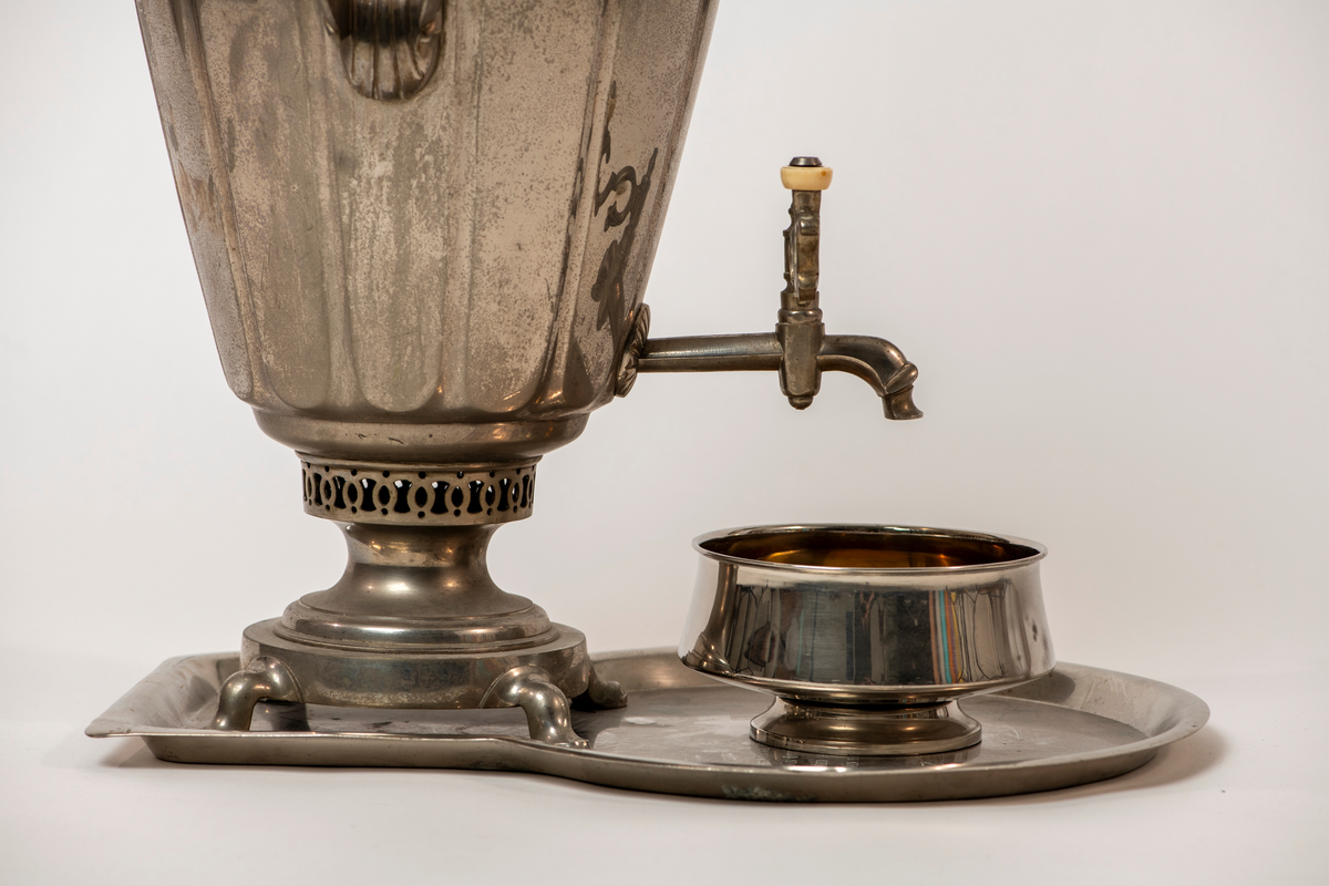 Samovar. Rund fot på fire utskrånende føtter. Stett med gjennombrutt dekor. Korpus med sider med rifler og avlange fasetter. To runde, tverr-riflede hanker av ben. Profilert overstykke med to benknotter. Ovalt fabrikkmerke med russisk tekst. Toppstykke med tre gjennombrutte ornamenter, nedsenket tungt lokk med ben-knott. Kran nederst på korpus, åpning i bunnen, og liten åpning for damp på overstykket.

Pipe. Forkrommet, skaft av messing og ben. Høy sylinderformet med utskrådd fotstykke, uten bunn, stjerthank.

Dryppskål av messing, utvendig forkrommet. Skål på stett. Midt i bunnen innvendig er det et ovalt firmamerke med russisk tekst.

Brett av forkrommet messing. Firkantet smal del, den annen halvdel bredere med rundet kontur.