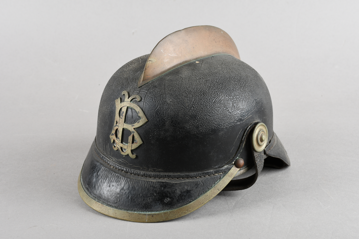 Brannhjelm med metallkam på toppen. Emblem i front/LB. Metallist rundt bremmen framme. Hakerem med feste i hver side, lukket med spenne. Skinnforing innvendig.