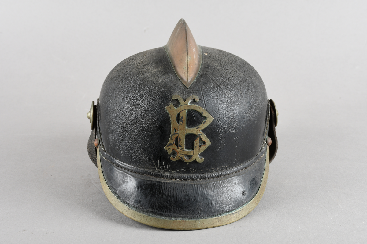 Brannhjelm med metallkam på toppen. Emblem i front/LB. Metallist rundt bremmen framme. Hakerem med feste i hver side, lukket med spenne. Skinnforing innvendig.