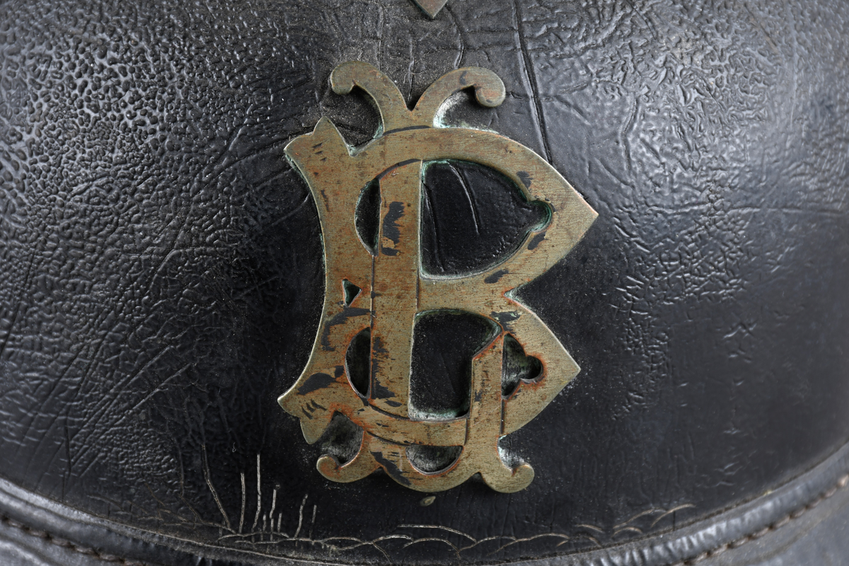 Brannhjelm med metallkam på toppen. Emblem i front/LB. Metallist rundt bremmen framme. Hakerem med feste i hver side, lukket med spenne. Skinnforing innvendig.