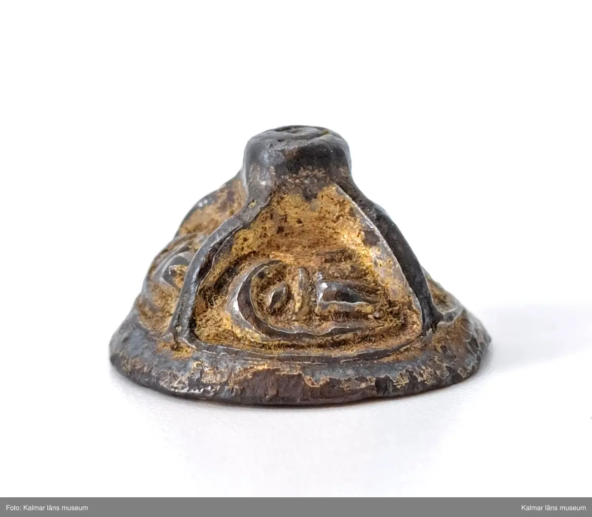 KLM 3048. Knapp. Gjuten del till en agraffknapp från folkvandringstid. Möjligen kan det också vara en svärdsknapp av samma typ  Liten, koniskt formad bronsknapp med förgyllning, som möjligen kan ha hört till en fibula. Översidan är uppdelad i fyra lika stora partier, i vardera ett stiliserat djurhuvud. Längst upp en knopp som är plan på ovansidan.