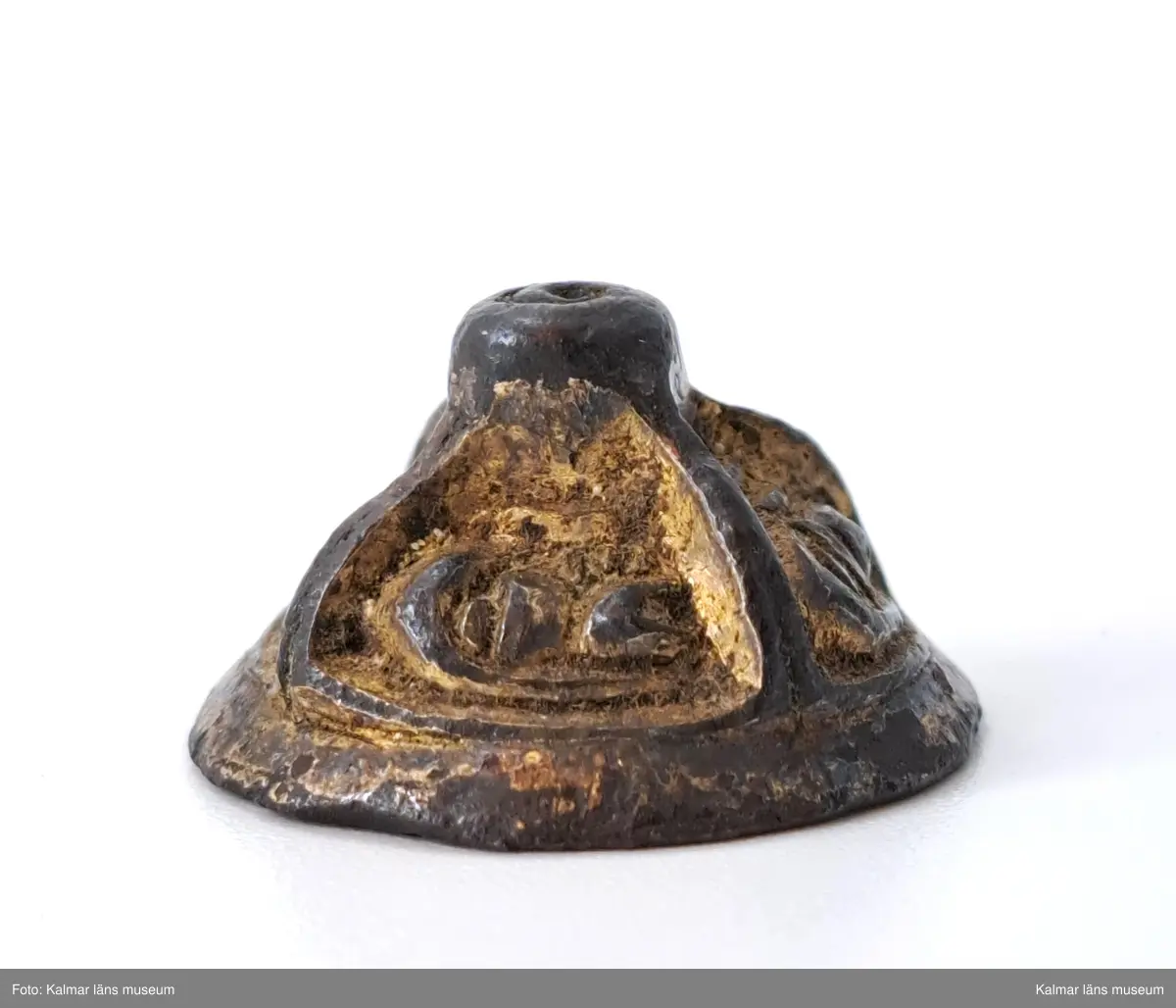 KLM 3048. Knapp. Gjuten del till en agraffknapp från folkvandringstid. Möjligen kan det också vara en svärdsknapp av samma typ  Liten, koniskt formad bronsknapp med förgyllning, som möjligen kan ha hört till en fibula. Översidan är uppdelad i fyra lika stora partier, i vardera ett stiliserat djurhuvud. Längst upp en knopp som är plan på ovansidan.