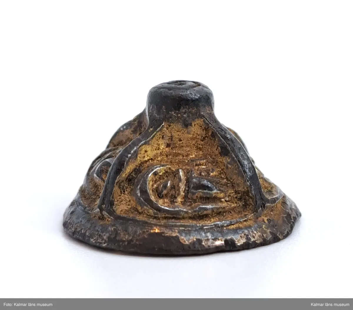 KLM 3048. Knapp. Gjuten del till en agraffknapp från folkvandringstid. Möjligen kan det också vara en svärdsknapp av samma typ  Liten, koniskt formad bronsknapp med förgyllning, som möjligen kan ha hört till en fibula. Översidan är uppdelad i fyra lika stora partier, i vardera ett stiliserat djurhuvud. Längst upp en knopp som är plan på ovansidan.