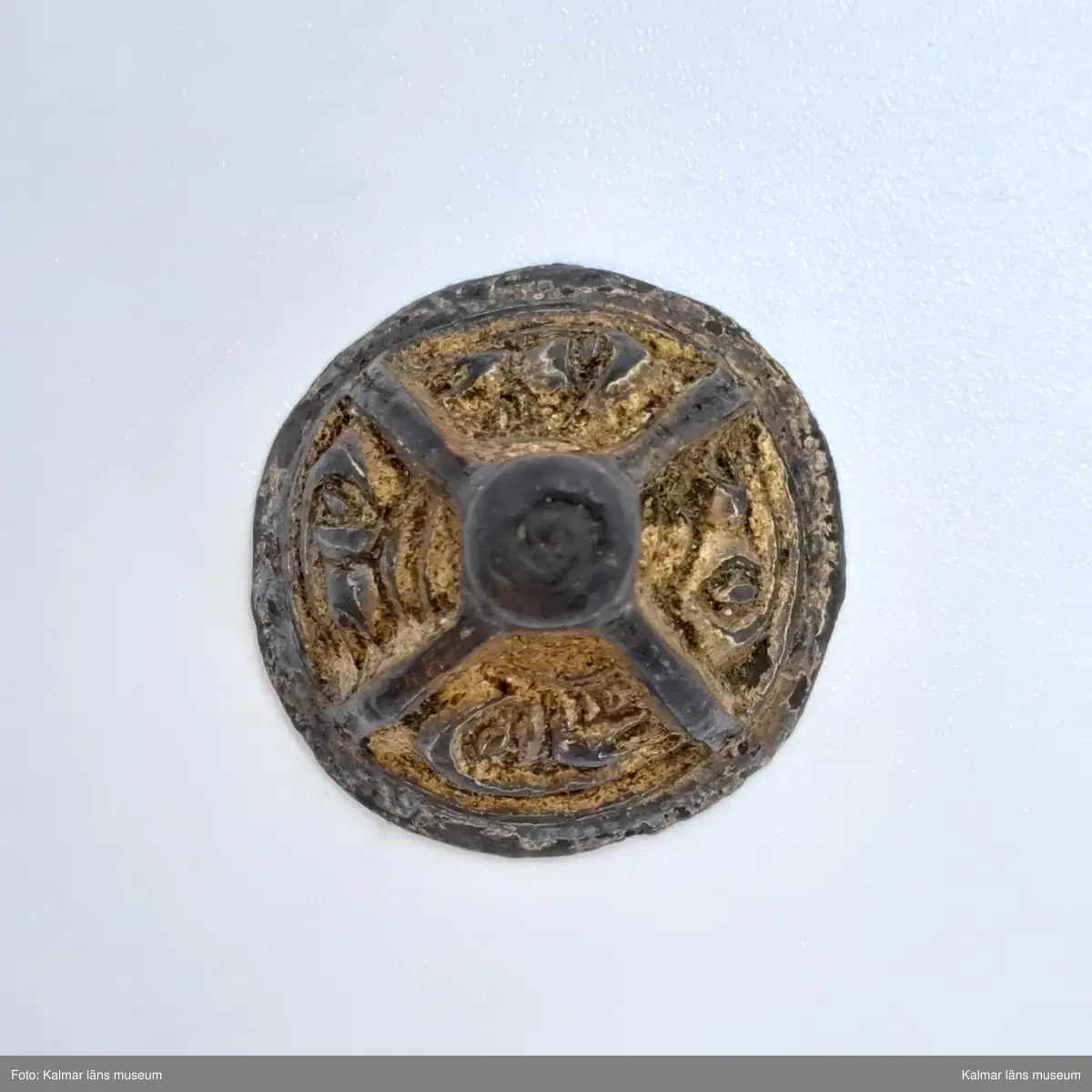 KLM 3048. Knapp. Gjuten del till en agraffknapp från folkvandringstid. Möjligen kan det också vara en svärdsknapp av samma typ  Liten, koniskt formad bronsknapp med förgyllning, som möjligen kan ha hört till en fibula. Översidan är uppdelad i fyra lika stora partier, i vardera ett stiliserat djurhuvud. Längst upp en knopp som är plan på ovansidan.