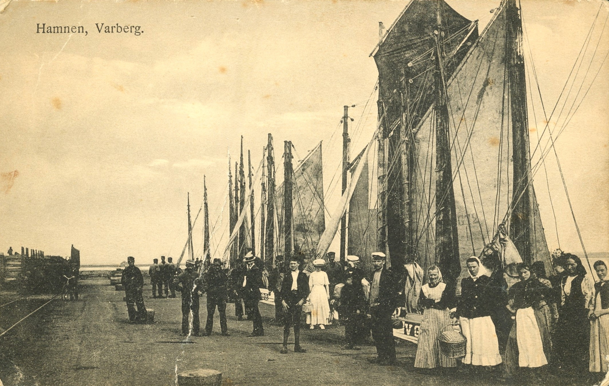 Vykort, "Hamnen, Varberg." En folksamling, vänd mot fotografen, vid fiskebåtarna längs västra kajen i Varbergs hamn. Tidigt 1900-tal. Bild 2 är en svartvit version.