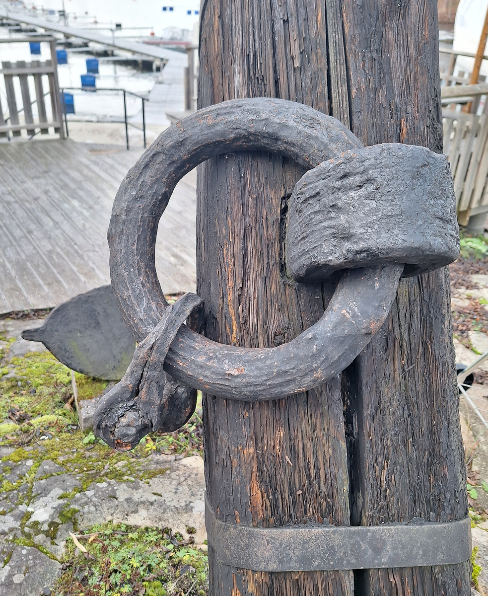 Stockankare. Trästock med fyra järnband samt en skyddskåpa i metall över ändträet på övre delen. Stockens två delar hålls ihop med fyra kilade trädymlingar. Stocken är på vissa partier mycket sliten. Schackel i röringen.