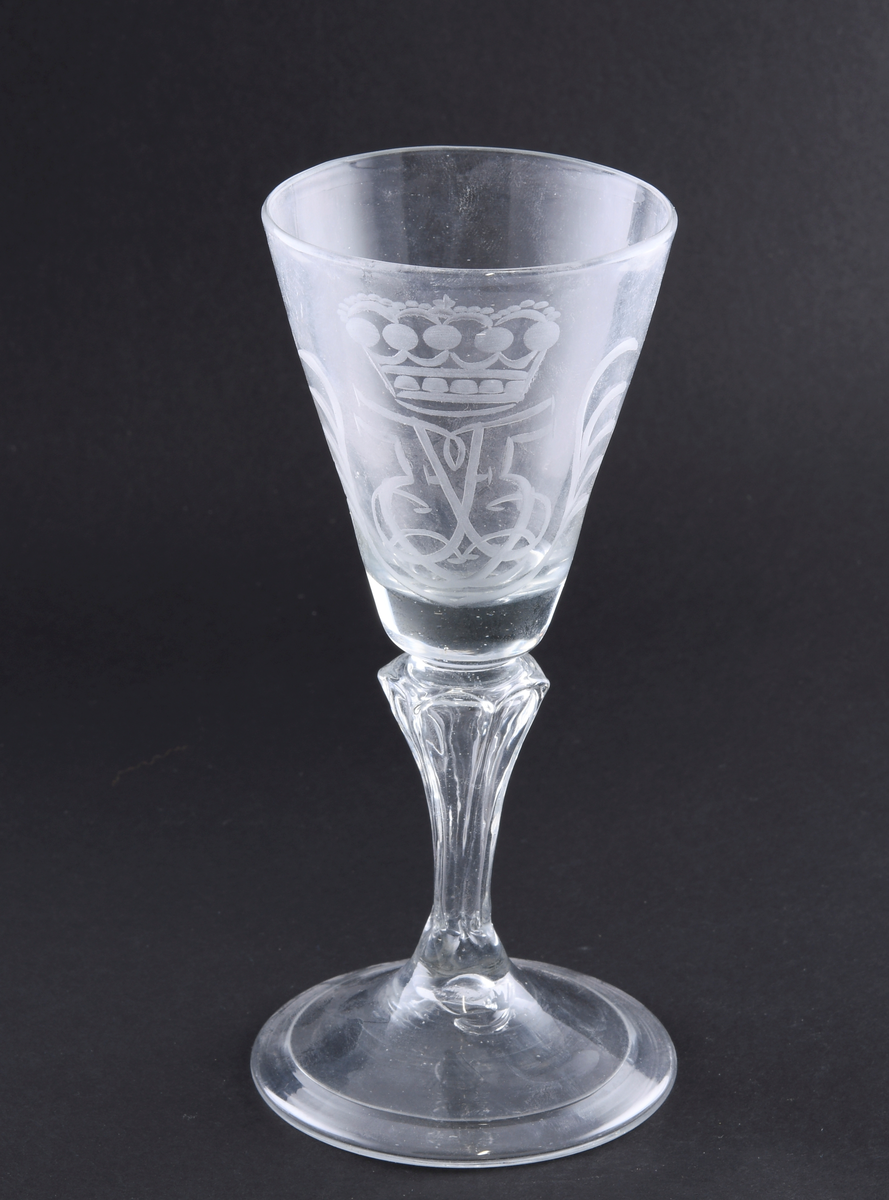 Vinglass med konisk klokke. På klokken er det gravert dekor med motiv av monogrammet til kong Fredrik V. Stetten er diamantfasettert. Foten er rund, med dekorativ innfelling ved randen.