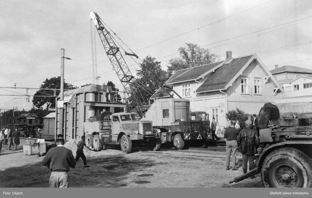 Transport av transformator fra Ise stasjon til Hasle i Varteig juli 1963. Bildet er tatt på Ise jernbanestasjon. Boggien, flerakslet bakstell på lastebiler og tilhengere (trailere) veide 46 tonn. Kranbil løfter over transformatoren fra jernbanevogn. Tilskuere. Husene bak til høyre er Knut Titterud pølsemakeri & forretning.
Lastebilen er en Diamond T 980. Bilkjennetegn A-1314.