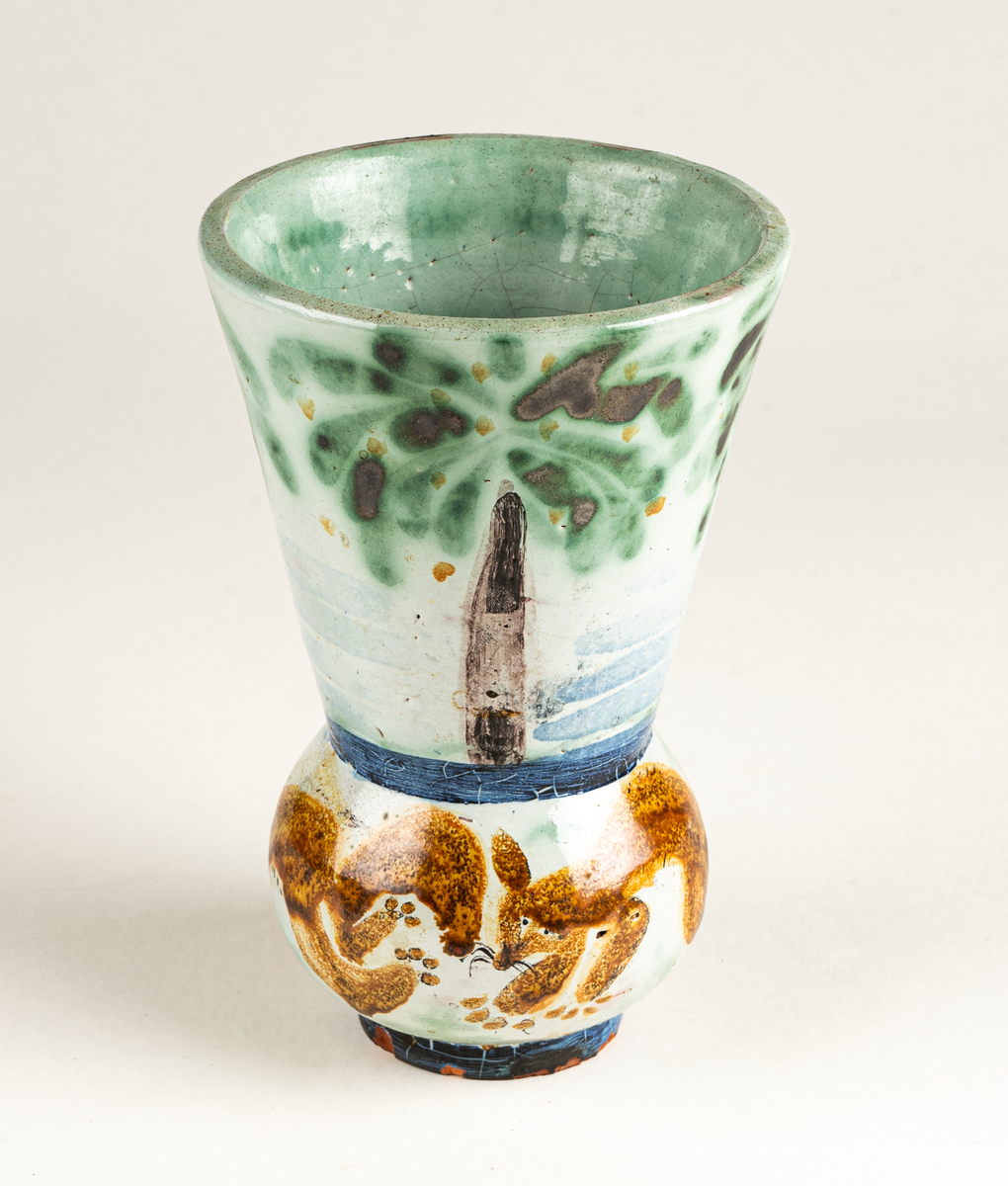 Vas, övre delen glaserad med trädmotiv i grönt, blått och brunt. Den nedre delens motiv föreställer springande ekorrar. Formgiven av Märta Willborg, 1930.