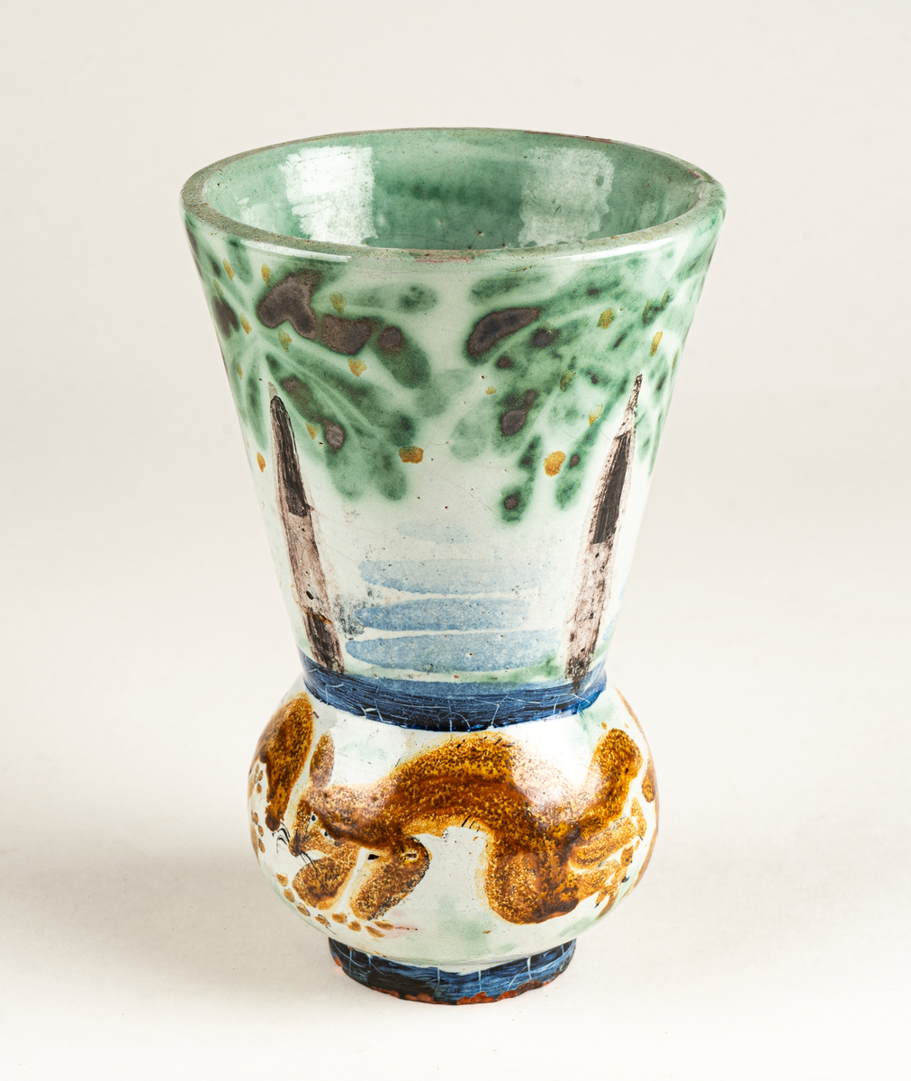 Vas, övre delen glaserad med trädmotiv i grönt, blått och brunt. Den nedre delens motiv föreställer springande ekorrar. Formgiven av Märta Willborg, 1930.