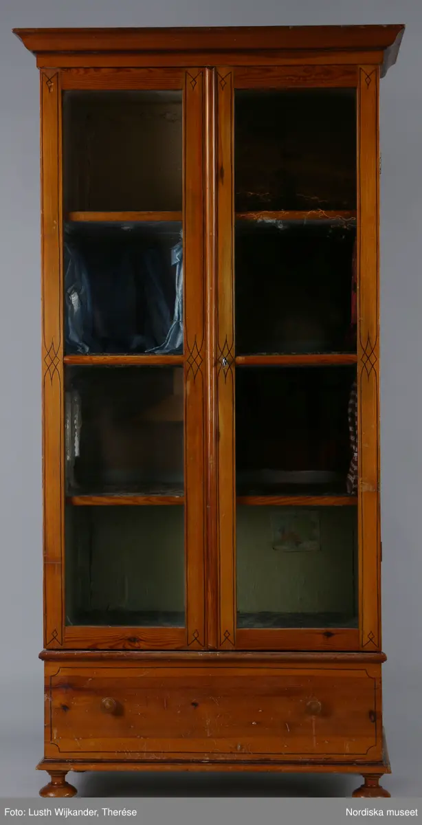 Inventering Sesam 1996-1999:
L 79 cm, B 46 cm, H 174 cm
Dockskåp av brunbetsat furu. Skåp med ett par glasdörrar med brun dekorrand på ramarna, undertill en låda med två dragknoppar, rakt krön med profilerad list. Skåpet står på fyra kulfötter. Dörrparet med lås och nyckel. På gavlarna 4 st fönster på vardera sidan. I skåpet 4 st våningar med sammanlagt 7 rum. Samtliga rum med mönstrade papperstapeter och krok i taket för lampa.
VÅNING 1: 1 rum, heltäckningsmatta av blåmönstrat tyg på vit botten. På höger kortvägg 3 st små pappersbilder i färgtryckav liten flicka i olika situationer. På  bakväggen en europakarta med tysk text.
VÅNING 2:  2 st rum. Bägge med mönstrade linoleummattor och två fönster. Gråmålad dörr mellan rummen. Vänster rum; Spiskåpa av vit papp på bakväggen. 2 st gardinlufter av vit spets med kornischer av gulmetall med påklistrad guldpappersremsa med mönster i relief. Höger rum; två gardinlufter av brunrandigt tyg, kornischer som ovan, omtag av metallkedjor på ena luften och rosa sidenband på andra. Ovanför dörren sitter 3 st beslag av pressad plåt. En större med måne och blommor och två mindre med sköld och blommor. 
VÅNING 3: bägge rummen med mönstrad heltäckningsmatta med lugg och två fönster. Vänster rum; 2 gardinlufter, 1 sänghimmel och 1 draperi av blått siden med vit kantspets. Gardiner med kornischer som ovan. 5 st omtag i form av snodd av gulmetall. Höger rum; enfärgade röda tapeter,2 st gardinlufter av rödrandigt siden med omtag av samma tyg, undergardin av vit spets, kornischer som ovan. Ett draperi av rött och randigt (i flera färger) ylle med ett omtag av metallkedja.
VÅNING 4: Vänster rum; flerfärgat och småmönstrad linoleummatta, Höger rum; utan golvbeläggning. 
- Ett par gröna gardiner av stärkt bomull, som ursprunglingen har suttit innanför glasdörrarna på dockskåpet. Rynkade i båda kortändorna. B 46,5, L 128 cm
Har tillhört givaren. (född 1886 enl. adelskalendern). 
Inventarier till dockskåpet inv.nr 164571-164728
Leif Wallin nov 1997