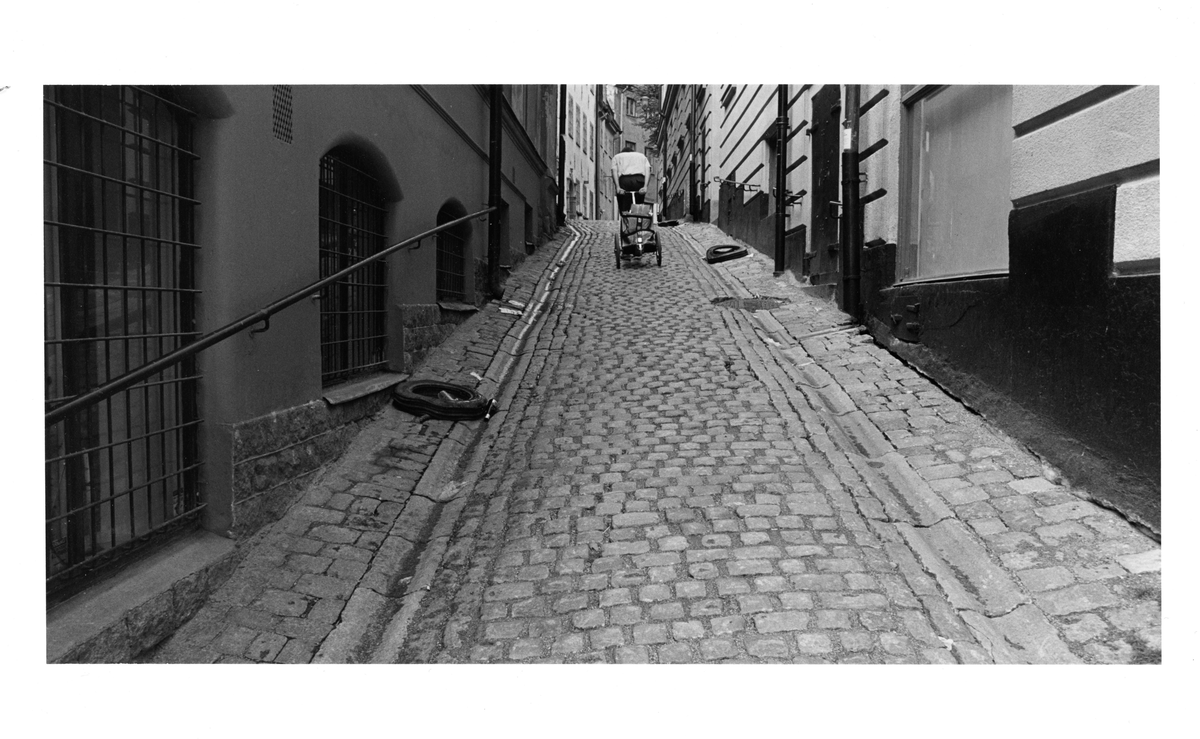 Brevbärare med brevbärarkärra gående uppför kullerstensgata.