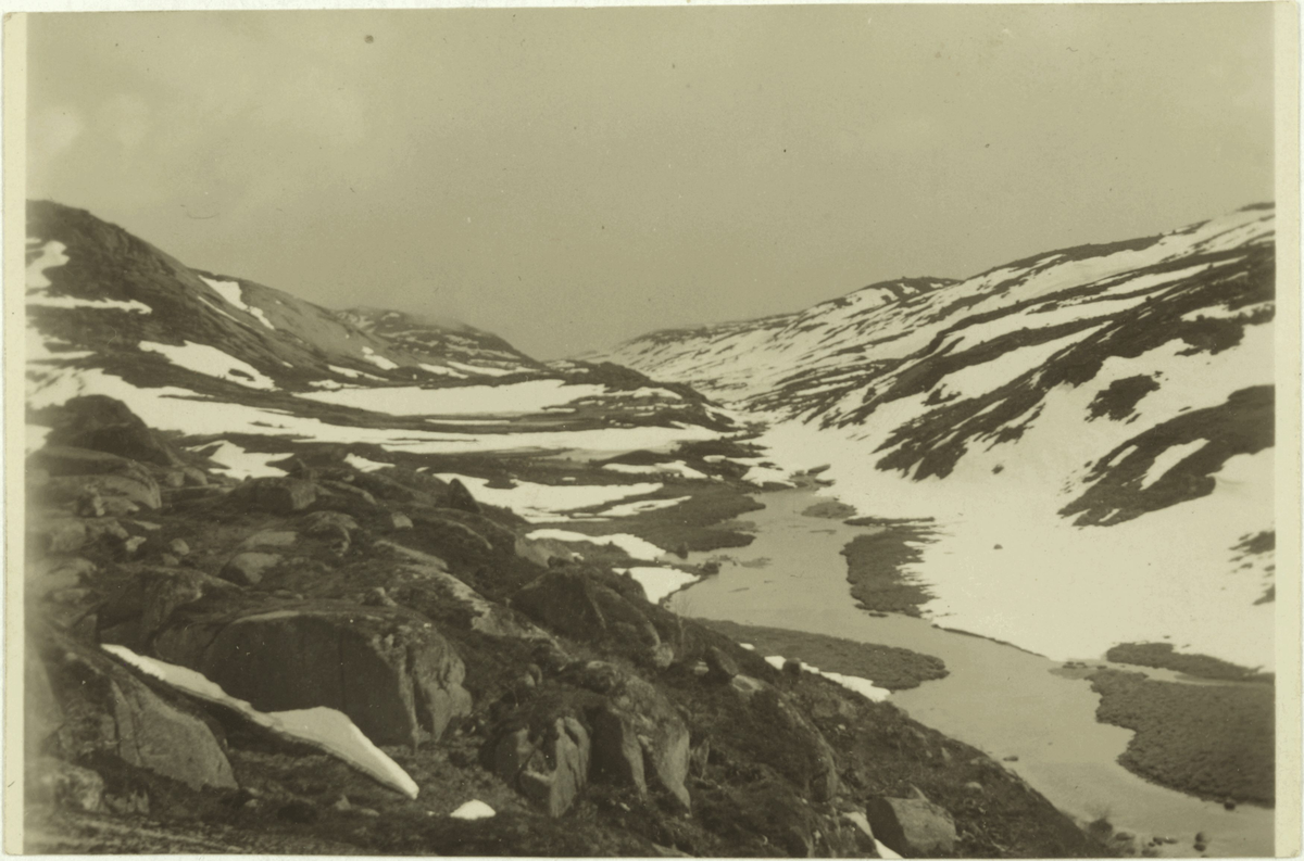 Eit snødekt fjellandskap med ei elv. På baksida av bildet står det: "ca. 900 m over havet den 1ste juni 1936. Jogledalen?".