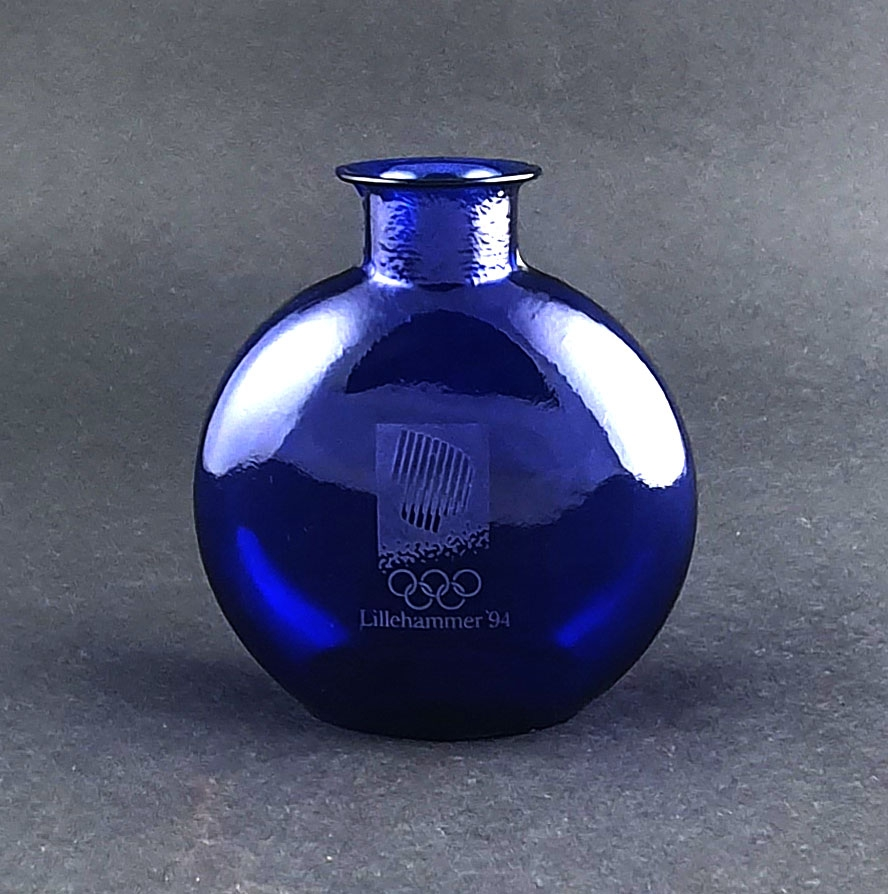 Blå glassflaske med rund korpus. På den ene siden emblemet for Lillehammer '94, og på den andre siden piktogrammet for kunstløp.