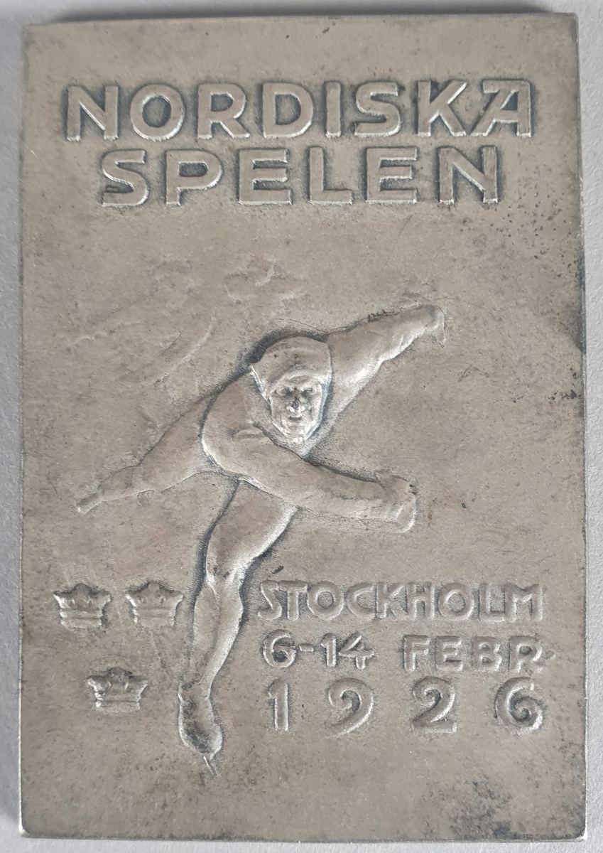 Firkantet medalje, rekangulær form, av sølv, med skøyteløpere og tre kroner i relieff.