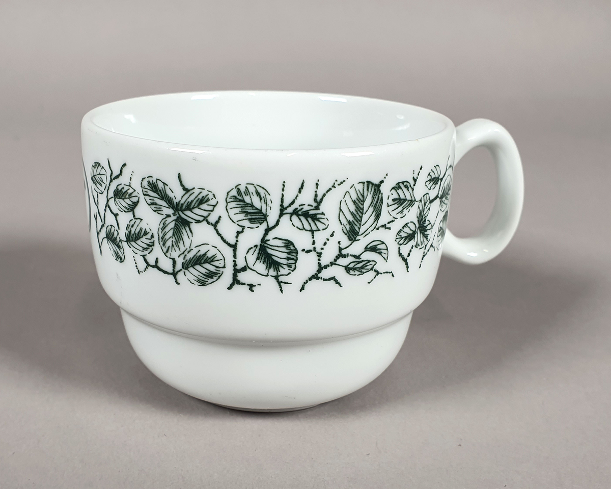 Hvit kaffekopp av porselen, med grønt bladmønster langs kanten og turistforeningens logo fra perioden etter 1988.
