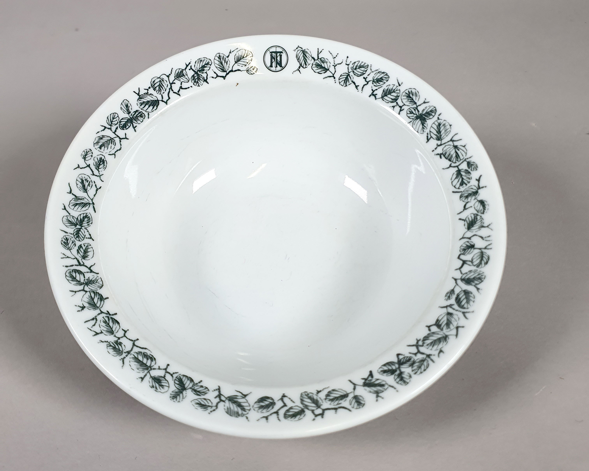 Hvit dyp tallerken av porselen, med grønt bladmønster langs kanten, og turistforeningens logo fra perioden 1943-1987.