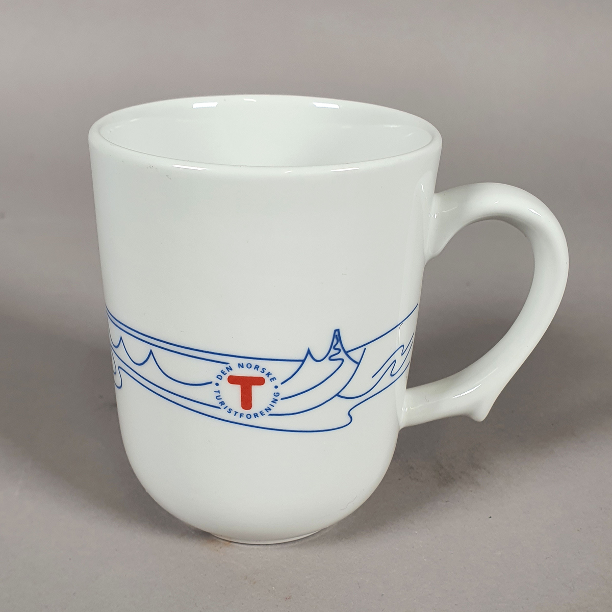 Hvit kopp av porselen, med med blått dekor (fjellmotiv) rundt korpus, og turistforeningens logo fra 1988