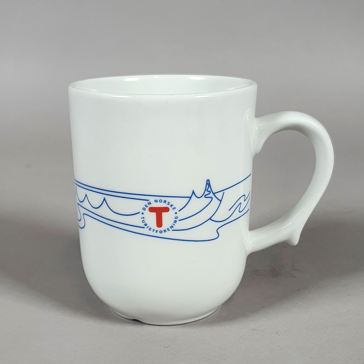 Hvit kopp av porselen, med med blått dekor (fjellmotiv) rundt korpus, og turistforeningens logo fra 1988
