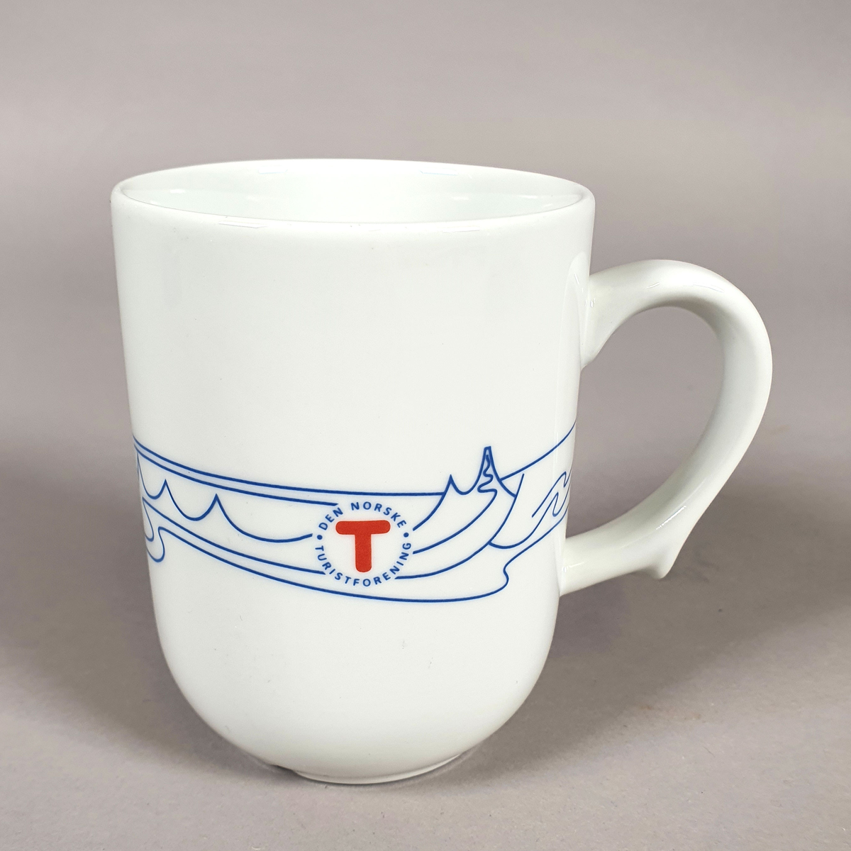 Hvit kopp av porselen, med med blått dekor (fjellmotiv) rundt korpus, og turistforeningens logo fra 1988
