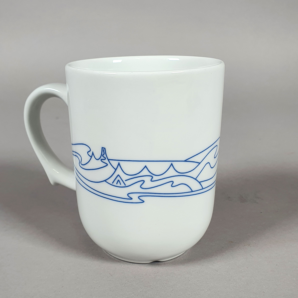 Hvit kopp av porselen, med med blått dekor (fjellmotiv) rundt korpus, og turistforeningens logo fra 1988
