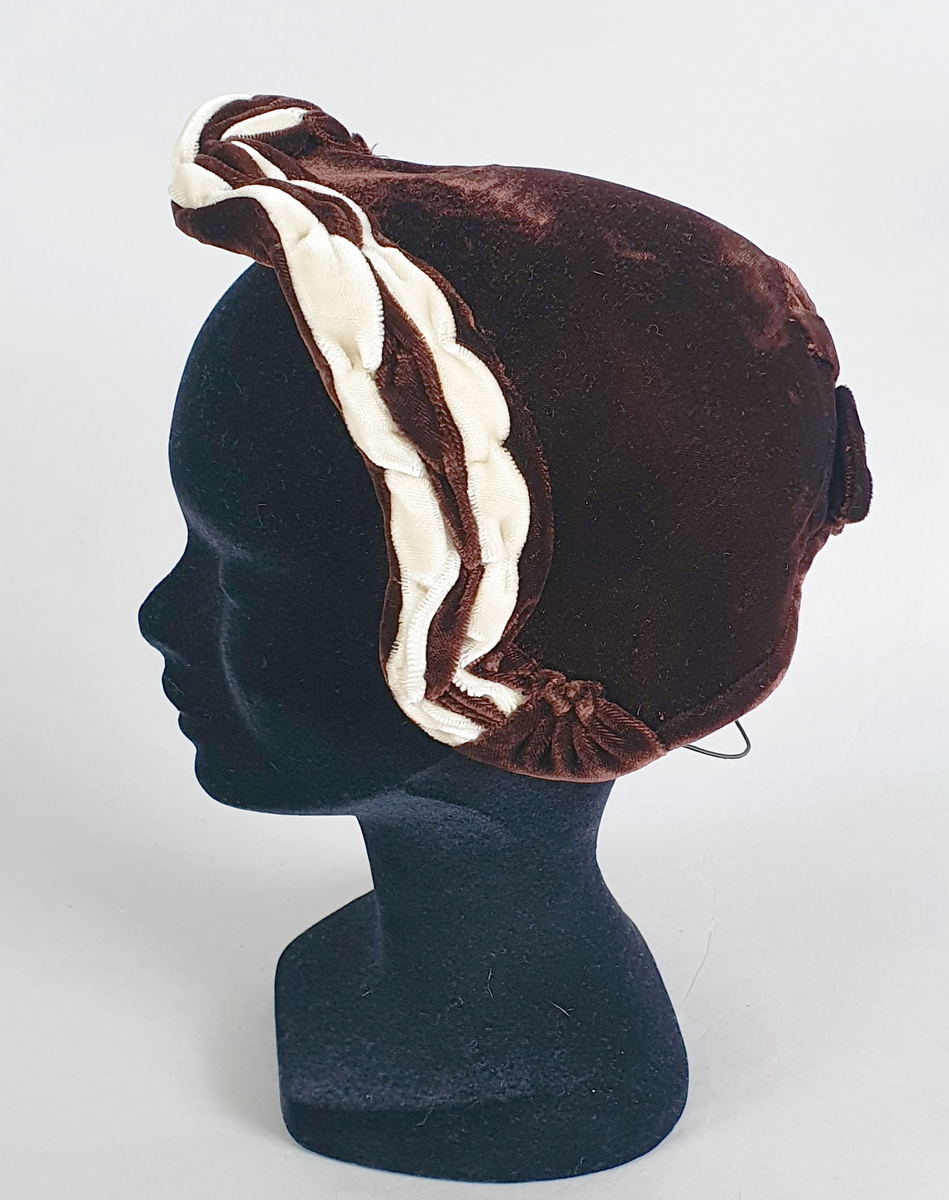 Brun damehatt av fløyels med hvitt dekor langs kanten foran og brun fløyelssløyfe midt bak. Spiler på innsiden langs kantene. Fôret med lysebrun kunstsilke og ripsbånd. Produsentmerke på innsiden.