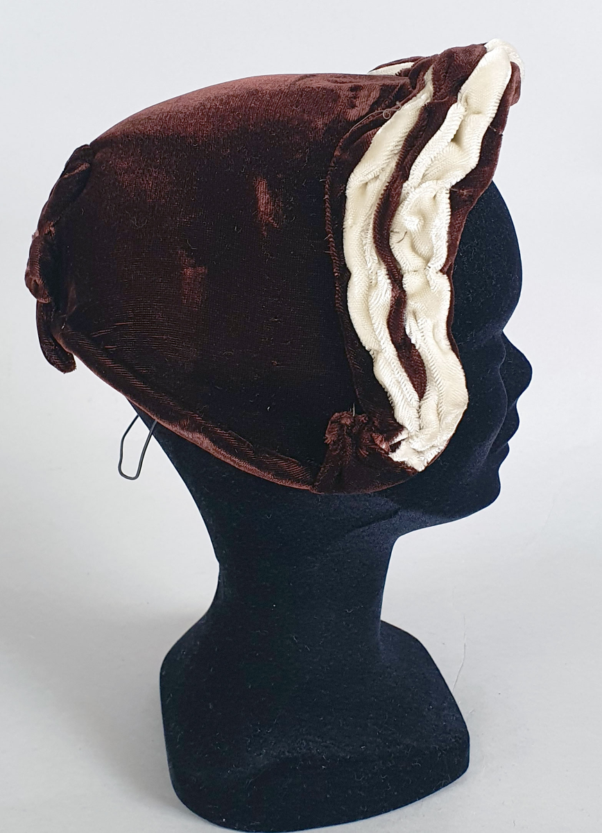 Brun damehatt av fløyels med hvitt dekor langs kanten foran og brun fløyelssløyfe midt bak. Spiler på innsiden langs kantene. Fôret med lysebrun kunstsilke og ripsbånd. Produsentmerke på innsiden.