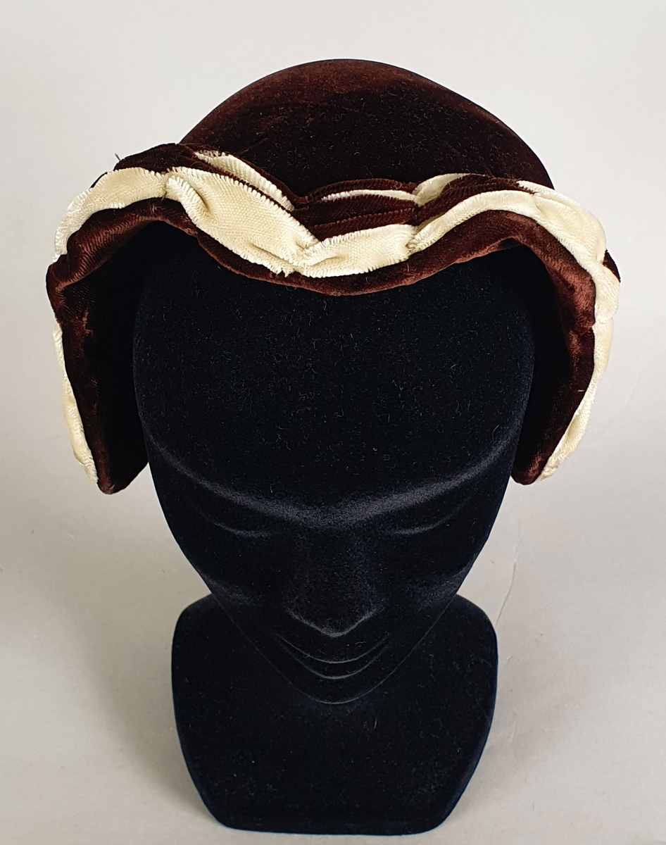 Brun damehatt av fløyels med hvitt dekor langs kanten foran og brun fløyelssløyfe midt bak. Spiler på innsiden langs kantene. Fôret med lysebrun kunstsilke og ripsbånd. Produsentmerke på innsiden.