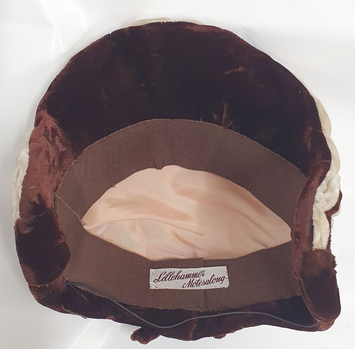 Brun damehatt av fløyels med hvitt dekor langs kanten foran og brun fløyelssløyfe midt bak. Spiler på innsiden langs kantene. Fôret med lysebrun kunstsilke og ripsbånd. Produsentmerke på innsiden.
