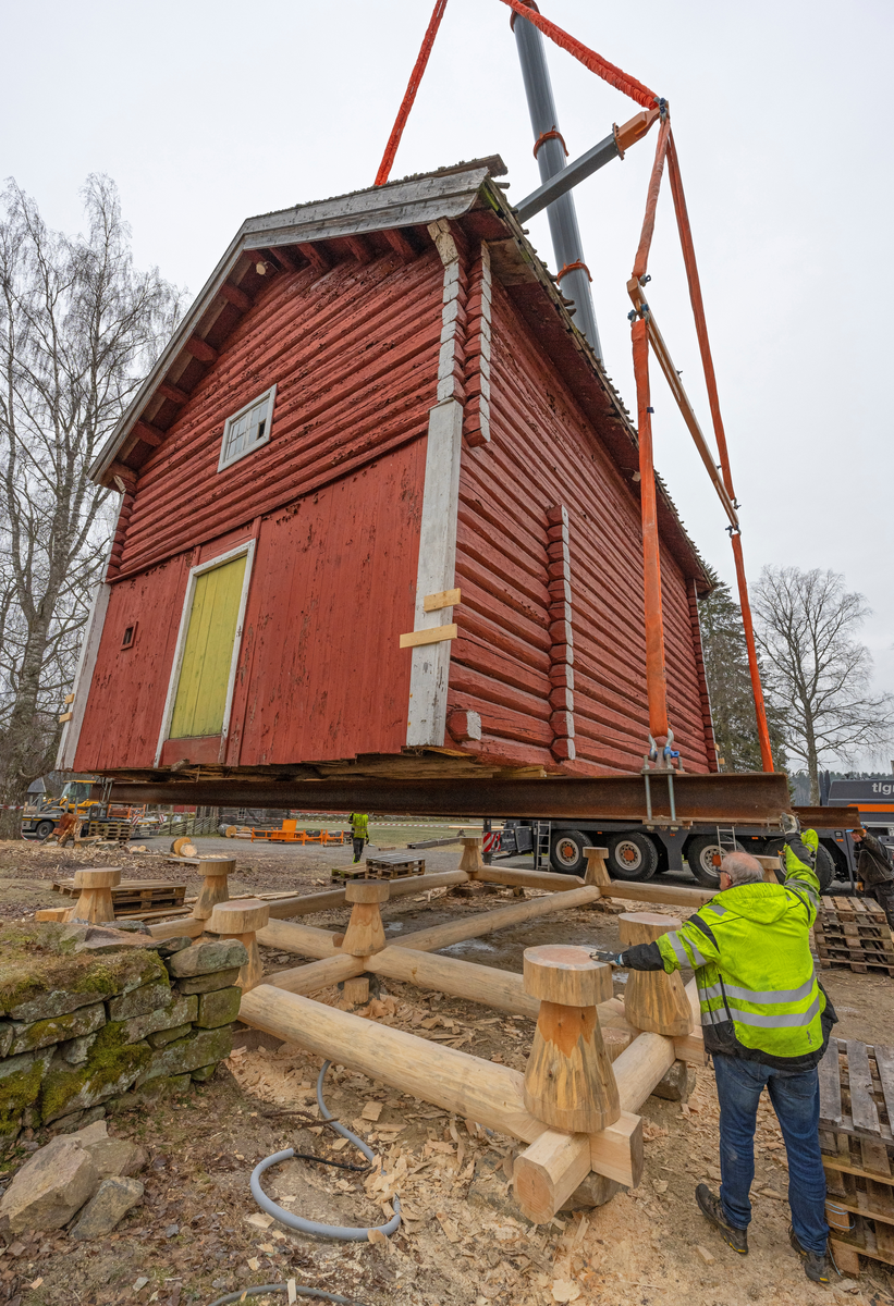 Heising av stabburet på klokkergarden Fossum på Norsk skogmuseum høsten 2024. Stabburet er en laftekonstruksjon som er om lag 8,4 meter langt, 6,2 meter bredt og har en rafthøyde på 4,4 meter. Det dreier seg med andre ord om en tung konstruksjon, og det hadde gjennom flere år bekymret personalet ved Norsk skogmuseum, som eier bygningen (jfr. SJF-B.0020), at den var i ferd med å synke ned i bakken. Dette førte til råteskader på svillramma og stabbene, særlig i den østre enden. Det var åpenbar fare for at underbygningen kunne kollapse, noe som kunne gitt alvorlige skader på «laftekassa». Tømrer Knut-Arild Nordli ble engasjert til å refundamentere bygningen. Den ble midlertidig løftet til side ved hjelp av kranbilen vi ser på dette bildet. Deretter ble det støpt betongsåler under bakkenivå som underlag for de ni laftesteinene som skulle bære den nye svillramma. På krysningspunktene i denne ramma – over de nevnte laftesteinene – ble det plassert ni cirka 70 centimeter høye stabber av furu med om lag 50 centimeters diameter. Deretter kunne bygningen løftes på plass igjen, slik vi ser det på dette bildet. Til høyre i forgrunnen står maskinentreprenør Arne Senderud, som på dette tidspunktet bisto med å få bygningen presist plassert på de nye fundamentene. Instrumentene til kranselskapet viste at bygningen veide drøyt 28 tonn.
