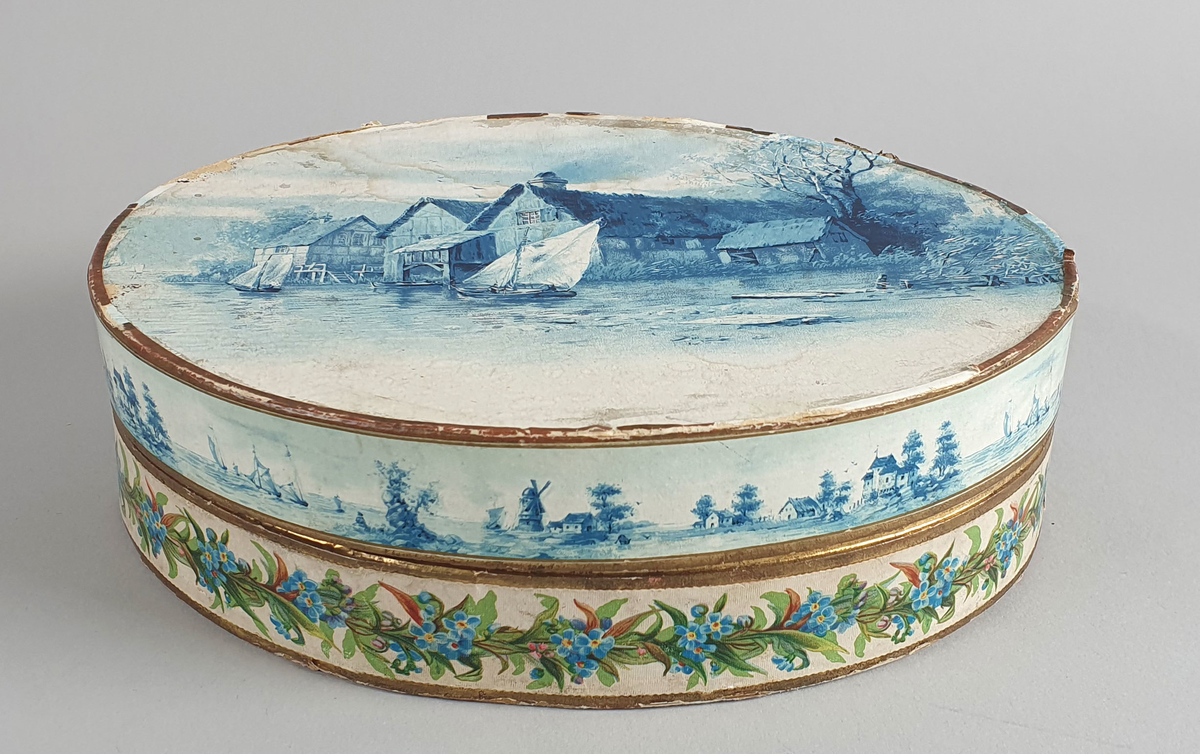 Oval eske med lokk. På lokket er det malt hus langs en elvebredd med båter i forgrunnen. Trær i bakgrunnen. I eske ligger det et nett av perler.  