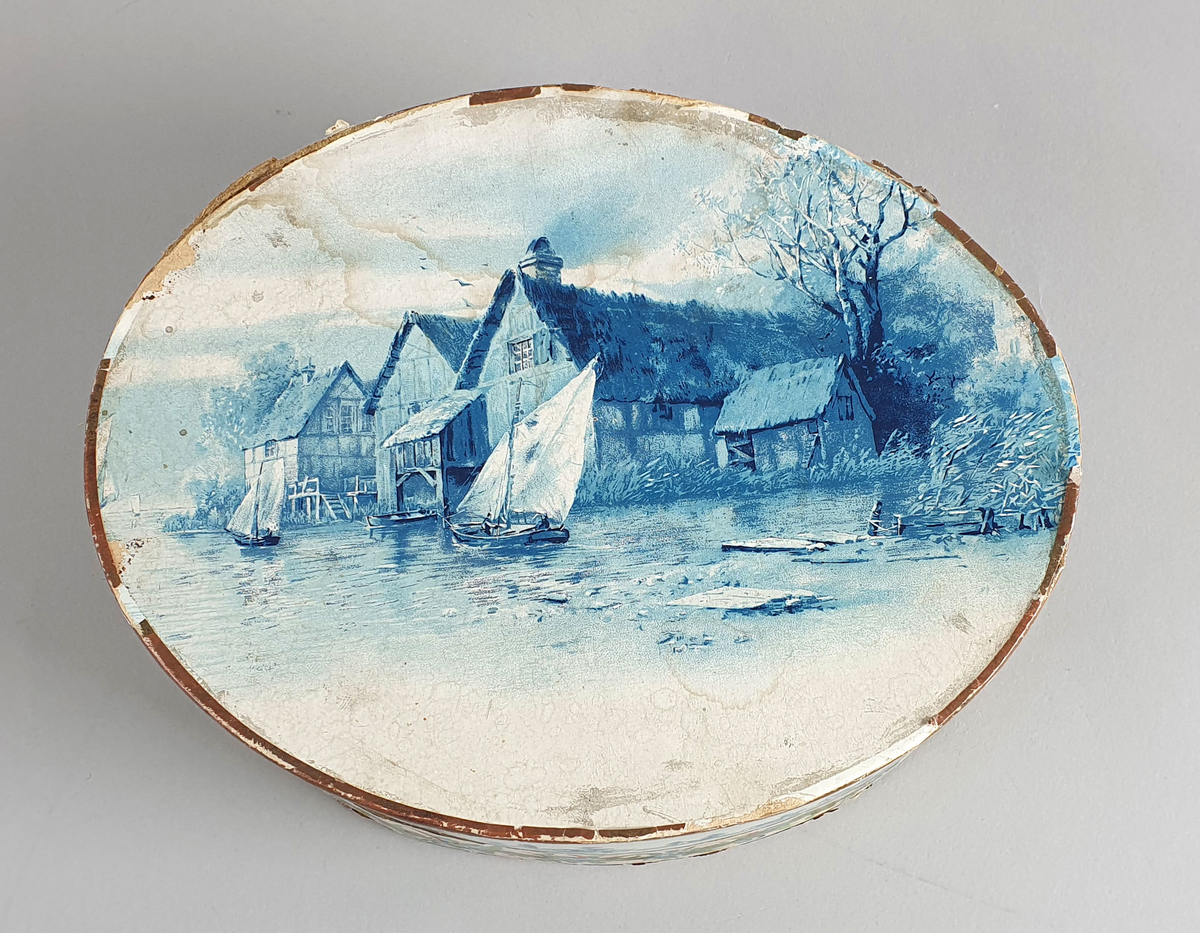 Oval eske med lokk. På lokket er det malt hus langs en elvebredd med båter i forgrunnen. Trær i bakgrunnen. I eske ligger det et nett av perler.  