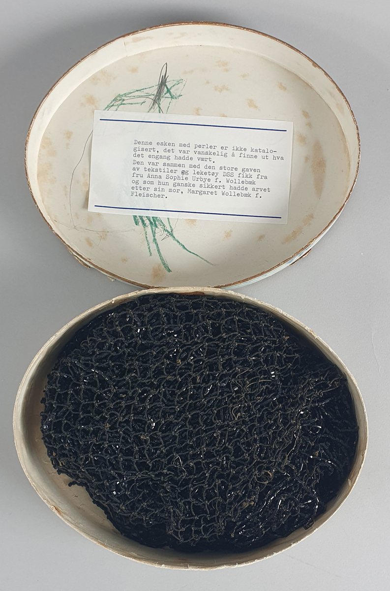 Oval eske med lokk. På lokket er det malt hus langs en elvebredd med båter i forgrunnen. Trær i bakgrunnen. I eske ligger det et nett av perler.  