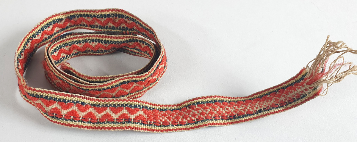 Smalt vevet bånd av ull med røde hjerter på naturfarget bunn. Grønne og gule striper langs kantene. 