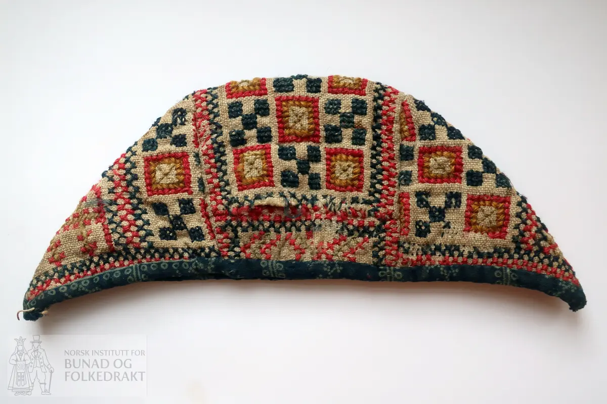 Barnehue. Trestykkshue med midtstykke og sidestykker. Midtstykket har ein skøyt på bakstykket, ca. 3 cm opp frå kanten. Hua er laga av lin med krosstingsbroderi i blått, raudt, lys grønt og gulbrunt over 3x3 trådar. Saumane inne i hua syner at dette er ombruk. Hua er kanta med blått bomullsstoff med lyseblått mønster, 0,6 cm breitt på retta og 0,9 cm på vranga, sydd på rette mot rette på retta, bretta over til vranga og sydd med kvit tråd, bomull? Fôr i grått bomullsstoff med små blomster i svart og kvitt.  Ytterstoff og fôr er sydd saman med ein saum 0,4 mm brei, bretta til kvar side og sydd ned. Ingen knyteband, men på eine sida ein liten rest av kvardingen, kanskje den er ein rest av eit knyteband?