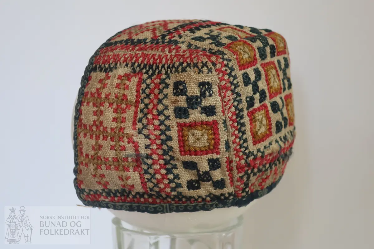 Barnehue. Trestykkshue med midtstykke og sidestykker. Midtstykket har ein skøyt på bakstykket, ca. 3 cm opp frå kanten. Hua er laga av lin med krosstingsbroderi i blått, raudt, lys grønt og gulbrunt over 3x3 trådar. Saumane inne i hua syner at dette er ombruk. Hua er kanta med blått bomullsstoff med lyseblått mønster, 0,6 cm breitt på retta og 0,9 cm på vranga, sydd på rette mot rette på retta, bretta over til vranga og sydd med kvit tråd, bomull? Fôr i grått bomullsstoff med små blomster i svart og kvitt.  Ytterstoff og fôr er sydd saman med ein saum 0,4 mm brei, bretta til kvar side og sydd ned. Ingen knyteband, men på eine sida ein liten rest av kvardingen, kanskje den er ein rest av eit knyteband?