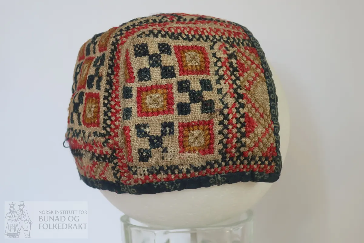 Barnehue. Trestykkshue med midtstykke og sidestykker. Midtstykket har ein skøyt på bakstykket, ca. 3 cm opp frå kanten. Hua er laga av lin med krosstingsbroderi i blått, raudt, lys grønt og gulbrunt over 3x3 trådar. Saumane inne i hua syner at dette er ombruk. Hua er kanta med blått bomullsstoff med lyseblått mønster, 0,6 cm breitt på retta og 0,9 cm på vranga, sydd på rette mot rette på retta, bretta over til vranga og sydd med kvit tråd, bomull? Fôr i grått bomullsstoff med små blomster i svart og kvitt.  Ytterstoff og fôr er sydd saman med ein saum 0,4 mm brei, bretta til kvar side og sydd ned. Ingen knyteband, men på eine sida ein liten rest av kvardingen, kanskje den er ein rest av eit knyteband?