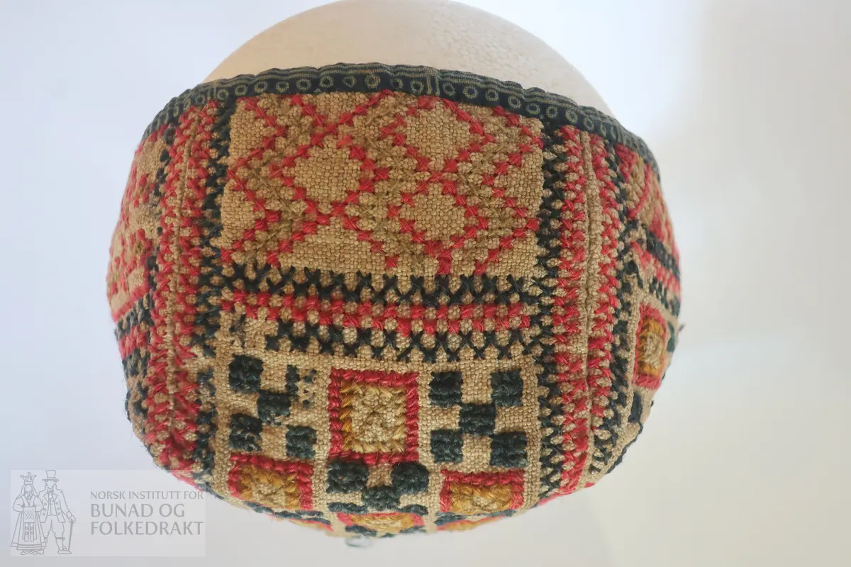 Barnehue. Trestykkshue med midtstykke og sidestykker. Midtstykket har ein skøyt på bakstykket, ca. 3 cm opp frå kanten. Hua er laga av lin med krosstingsbroderi i blått, raudt, lys grønt og gulbrunt over 3x3 trådar. Saumane inne i hua syner at dette er ombruk. Hua er kanta med blått bomullsstoff med lyseblått mønster, 0,6 cm breitt på retta og 0,9 cm på vranga, sydd på rette mot rette på retta, bretta over til vranga og sydd med kvit tråd, bomull? Fôr i grått bomullsstoff med små blomster i svart og kvitt.  Ytterstoff og fôr er sydd saman med ein saum 0,4 mm brei, bretta til kvar side og sydd ned. Ingen knyteband, men på eine sida ein liten rest av kvardingen, kanskje den er ein rest av eit knyteband?