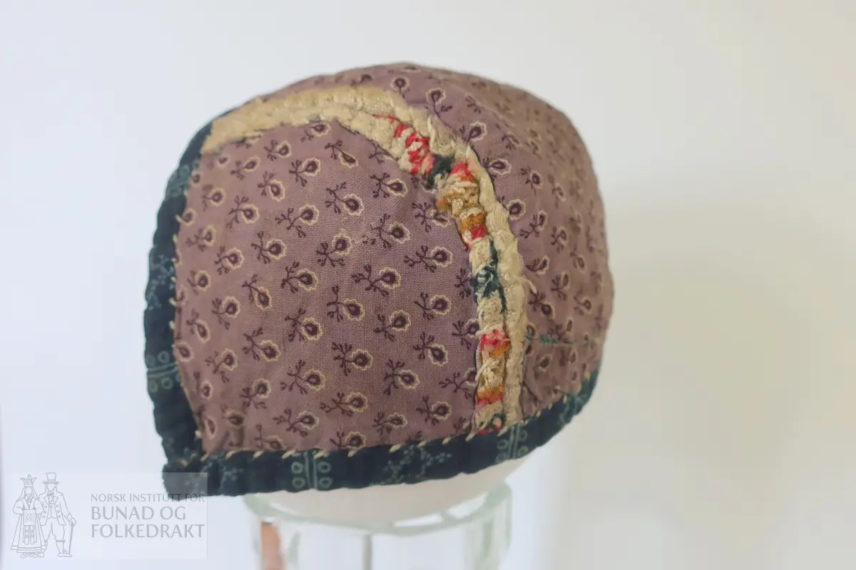 Barnehue. Trestykkshue med midtstykke og sidestykker. Midtstykket har ein skøyt på bakstykket, ca. 3 cm opp frå kanten. Hua er laga av lin med krosstingsbroderi i blått, raudt, lys grønt og gulbrunt over 3x3 trådar. Saumane inne i hua syner at dette er ombruk. Hua er kanta med blått bomullsstoff med lyseblått mønster, 0,6 cm breitt på retta og 0,9 cm på vranga, sydd på rette mot rette på retta, bretta over til vranga og sydd med kvit tråd, bomull? Fôr i grått bomullsstoff med små blomster i svart og kvitt.  Ytterstoff og fôr er sydd saman med ein saum 0,4 mm brei, bretta til kvar side og sydd ned. Ingen knyteband, men på eine sida ein liten rest av kvardingen, kanskje den er ein rest av eit knyteband?