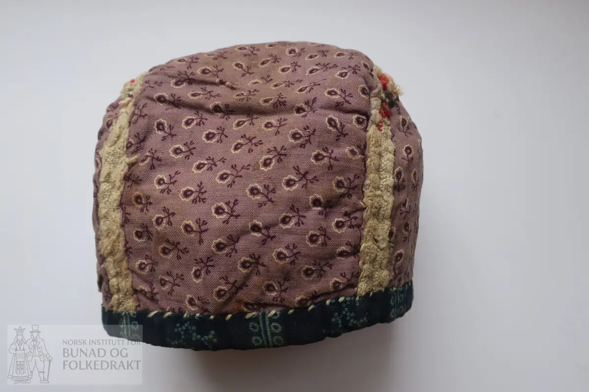 Barnehue. Trestykkshue med midtstykke og sidestykker. Midtstykket har ein skøyt på bakstykket, ca. 3 cm opp frå kanten. Hua er laga av lin med krosstingsbroderi i blått, raudt, lys grønt og gulbrunt over 3x3 trådar. Saumane inne i hua syner at dette er ombruk. Hua er kanta med blått bomullsstoff med lyseblått mønster, 0,6 cm breitt på retta og 0,9 cm på vranga, sydd på rette mot rette på retta, bretta over til vranga og sydd med kvit tråd, bomull? Fôr i grått bomullsstoff med små blomster i svart og kvitt.  Ytterstoff og fôr er sydd saman med ein saum 0,4 mm brei, bretta til kvar side og sydd ned. Ingen knyteband, men på eine sida ein liten rest av kvardingen, kanskje den er ein rest av eit knyteband?