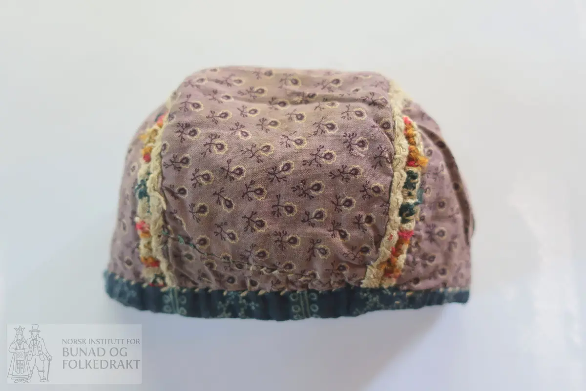 Barnehue. Trestykkshue med midtstykke og sidestykker. Midtstykket har ein skøyt på bakstykket, ca. 3 cm opp frå kanten. Hua er laga av lin med krosstingsbroderi i blått, raudt, lys grønt og gulbrunt over 3x3 trådar. Saumane inne i hua syner at dette er ombruk. Hua er kanta med blått bomullsstoff med lyseblått mønster, 0,6 cm breitt på retta og 0,9 cm på vranga, sydd på rette mot rette på retta, bretta over til vranga og sydd med kvit tråd, bomull? Fôr i grått bomullsstoff med små blomster i svart og kvitt.  Ytterstoff og fôr er sydd saman med ein saum 0,4 mm brei, bretta til kvar side og sydd ned. Ingen knyteband, men på eine sida ein liten rest av kvardingen, kanskje den er ein rest av eit knyteband?