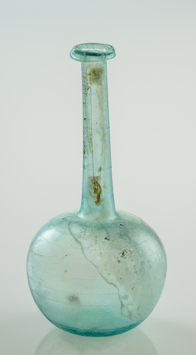 Flaska i blått glas. Bukig nedre del som övergår i en hög hals med utvikt mynningen. 
Slipade horizontella linjer över kupa och hals.