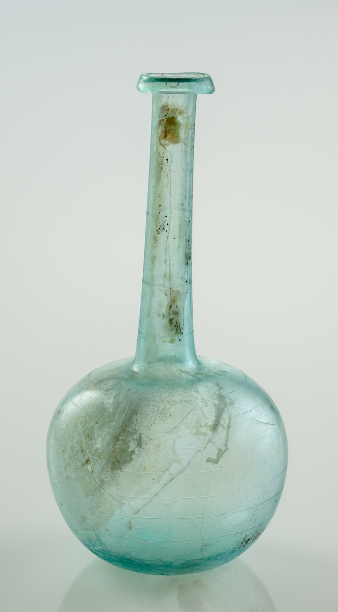 Flaska i blått glas. Bukig nedre del som övergår i en hög hals med utvikt mynningen. 
Slipade horizontella linjer över kupa och hals.